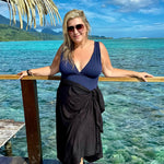 A person standing on a wooden deck wearing a black sarong, with the ocean and a tropical landscape in the background.