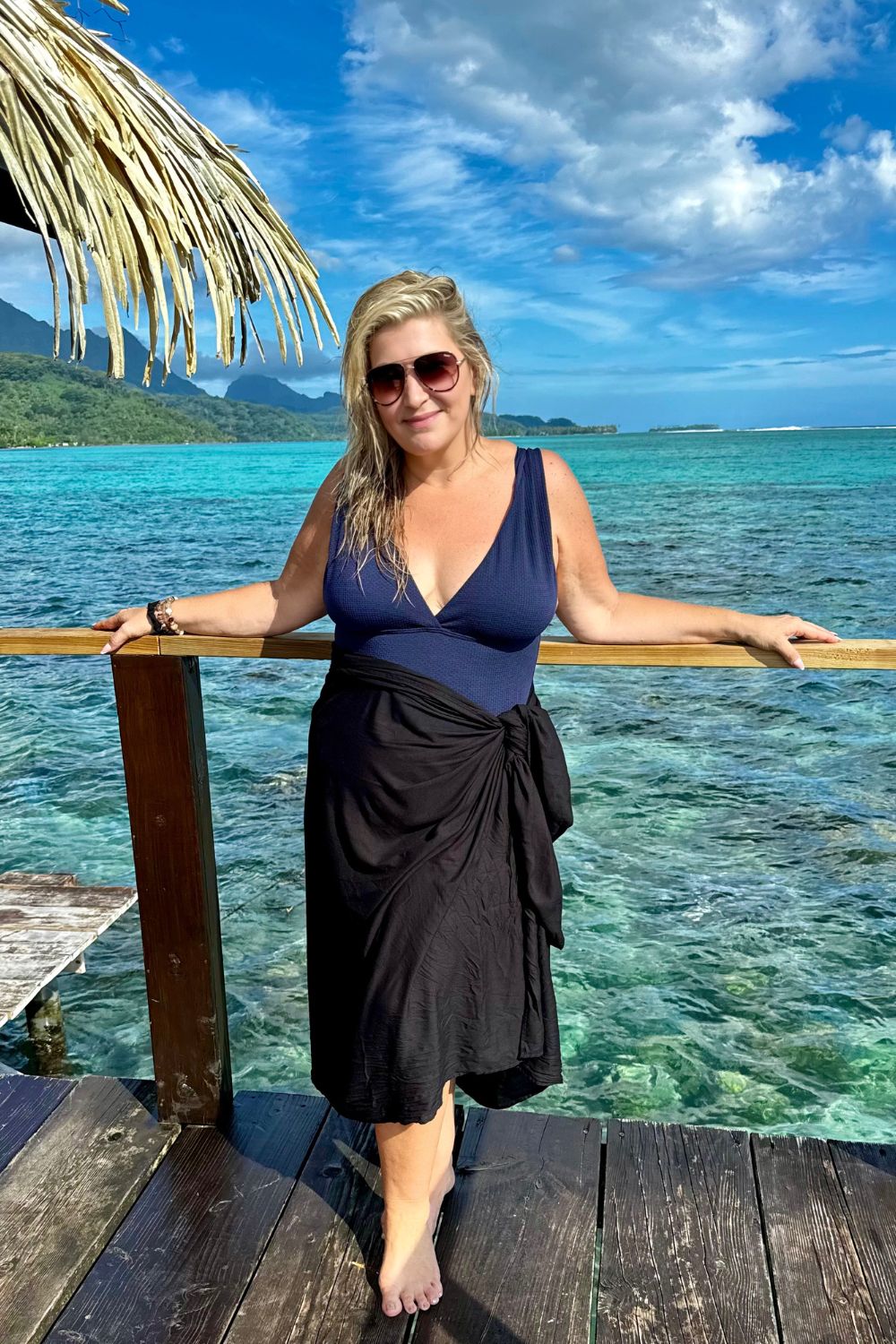 A person standing on a wooden deck wearing a black sarong, with the ocean and a tropical landscape in the background.