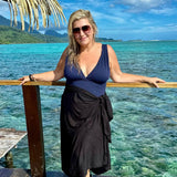 A person standing on a wooden deck wearing a black sarong, with the ocean and a tropical landscape in the background.