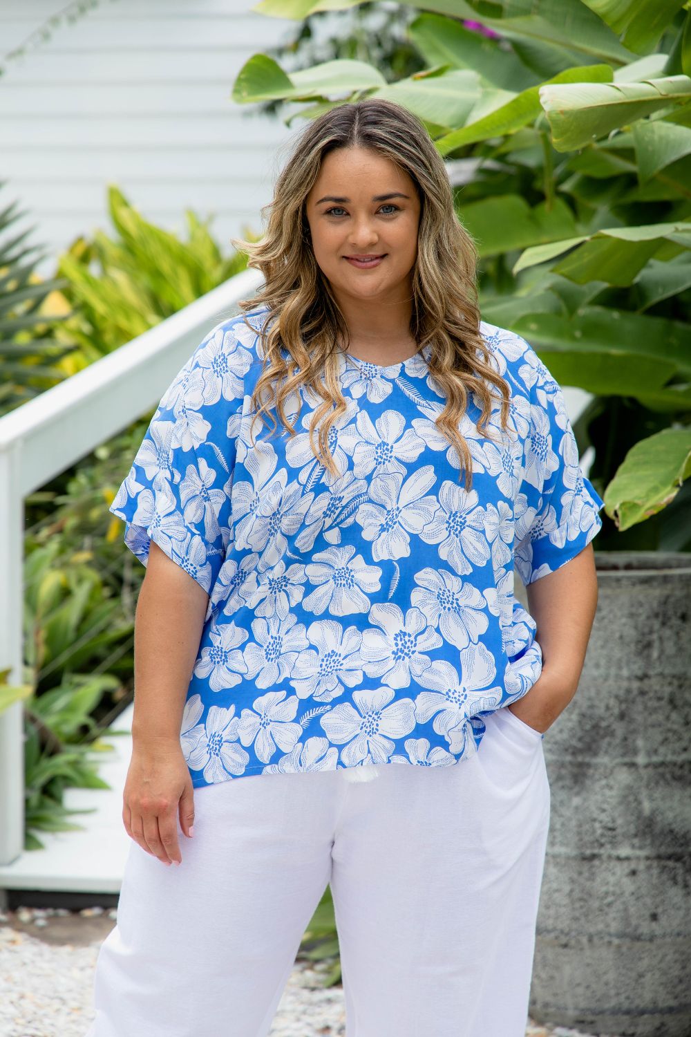 Woman wearing a blue floral top and white cotton pants standing outdoors with greenery in the background