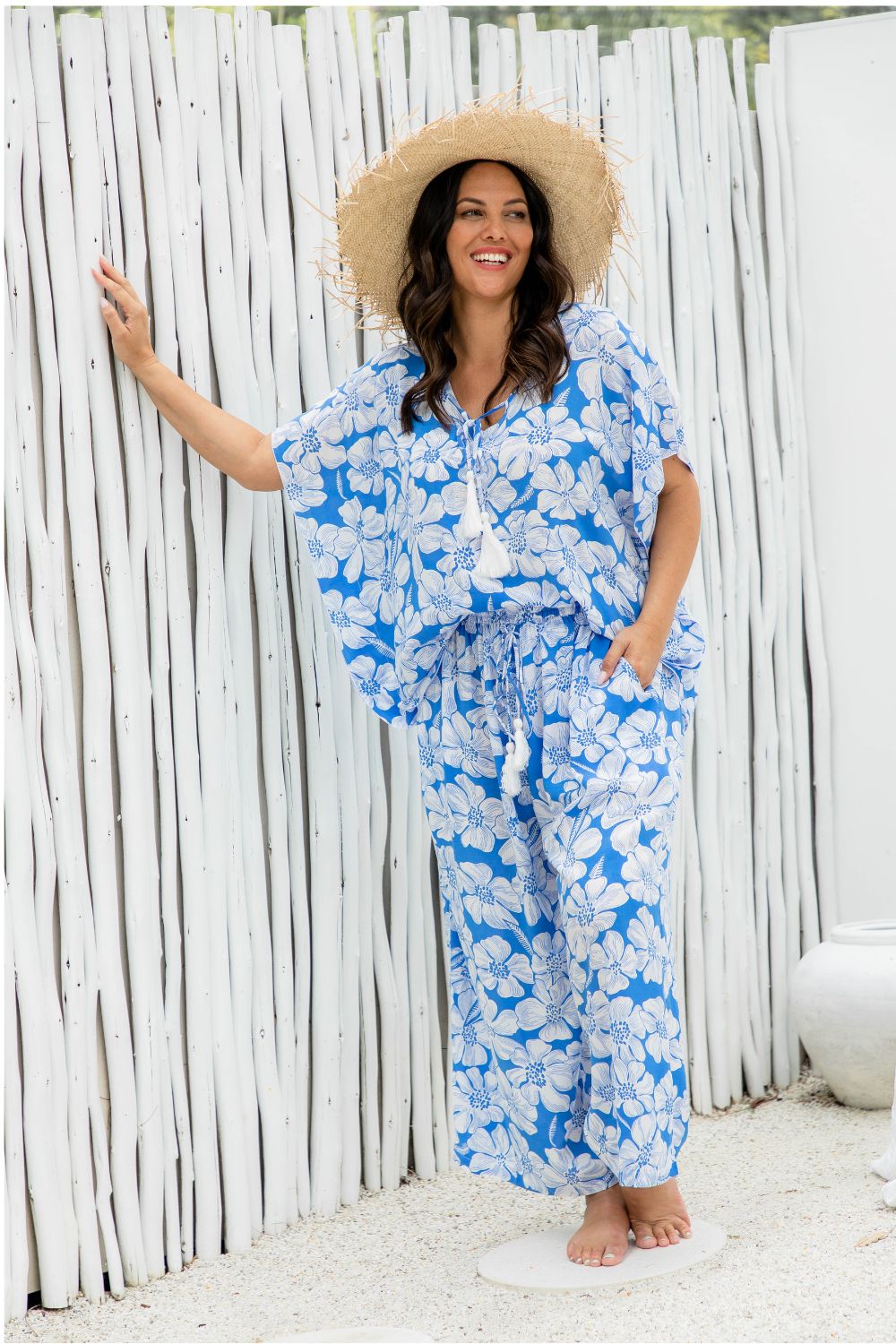 Woman wearing a blue floral pants with matching summer top against a white wooden fence of beach resort