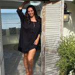 A woman wearing a black short-sleeve  short kaftan dress stands barefoot on a patio in front of an open doorway of a cafe in Thailand with shutters, with the ocean visible in the background.