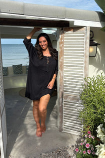 A woman wearing a black short-sleeve  short kaftan dress stands barefoot on a patio in front of an open doorway of a cafe in Thailand with shutters, with the ocean visible in the background.