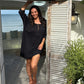A woman wearing a black short-sleeve  short kaftan dress stands barefoot on a patio in front of an open doorway of a cafe in Thailand with shutters, with the ocean visible in the background.