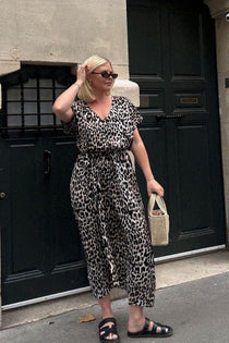 A woman wearing a leopard-print long kaftan dress, black sunglasses, and black slide sandals stands in front of a dark door, holding a small beige handbag whilst exploring the streets of Paris