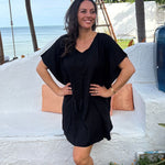 A woman wearing a black kimono sleeve kaftan dress stands outdoors in front of a white stucco wall with Thailand beach and palm trees in the background.
