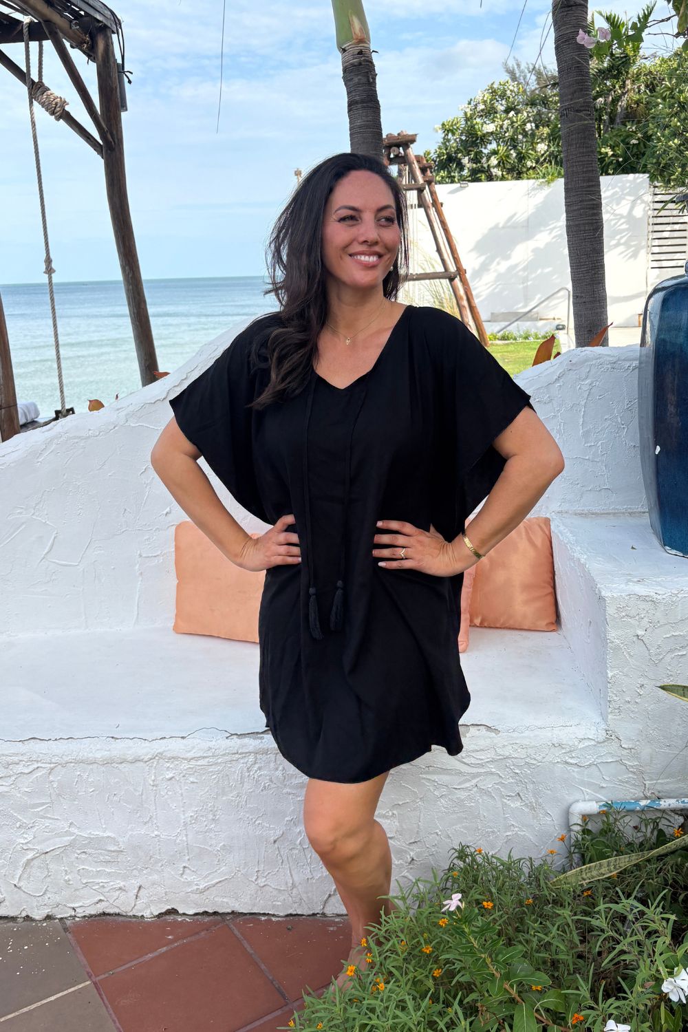 A woman wearing a black kimono sleeve kaftan dress stands outdoors in front of a white stucco wall with Thailand beach and palm trees in the background.