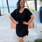 A woman wearing a black kimono sleeve kaftan dress stands outdoors in front of a white stucco wall with Thailand beach and palm trees in the background.
