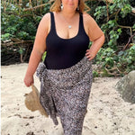 A woman stands on a sandy beach wearing a black swimmers and a black-brown and-white patterned sarong, in plus size, with tropical foliage and branches in the background.