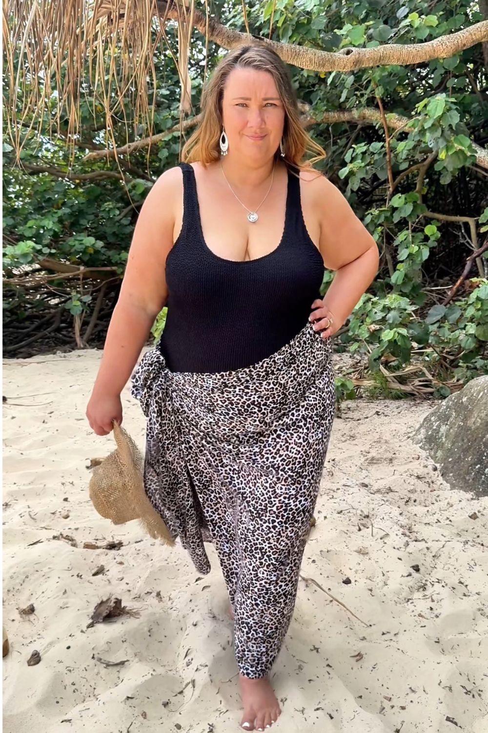 A woman stands on a sandy beach wearing a black swimmers and a black-brown and-white patterned sarong, in plus size, with tropical foliage and branches in the background.