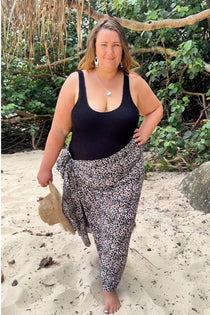 A woman stands on a sandy beach wearing a black swimmers and a black-brown and-white patterned sarong, in plus size, with tropical foliage and branches in the background.