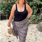 A woman stands on a sandy beach wearing a black swimmers and a black-brown and-white patterned sarong, in plus size, with tropical foliage and branches in the background.