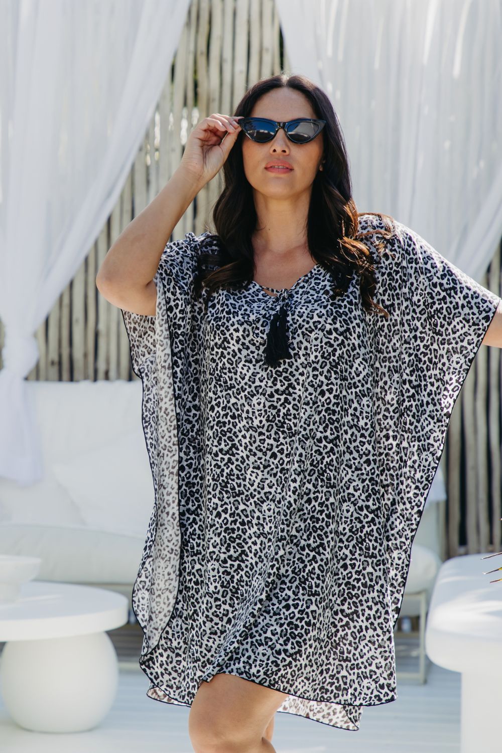 A woman wearing a black grey and white leopard print kaftan dress and sunglasses, posing in a modern outdoor lounge setting with white drapes and minimalist tables.