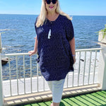 Mature age woman wearing the short kaftan dress in blue and white polka dot over white three quarter pants. Standing with the Italian coastline behind her whilst on summer holiday