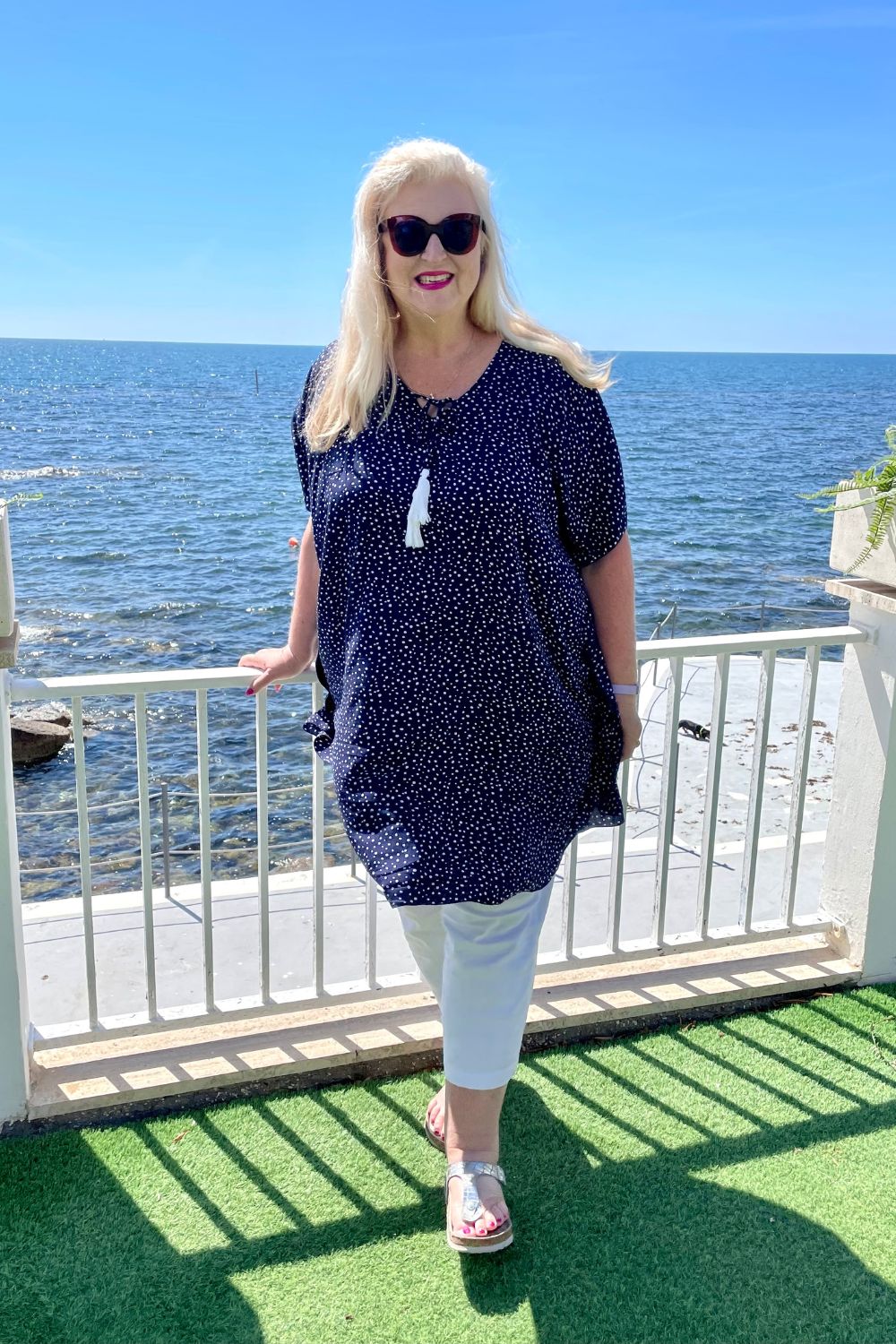 Mature age woman wearing the short kaftan dress in blue and white polka dot over white three quarter pants. Standing with the Italian coastline behind her whilst on summer holiday