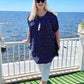 Mature age woman wearing the short kaftan dress in blue and white polka dot over white three quarter pants. Standing with the Italian coastline behind her whilst on summer holiday