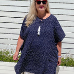 Mature age woman wearing the short kaftan dress in blue and white polka dot over white pants with planter boxes in background. 