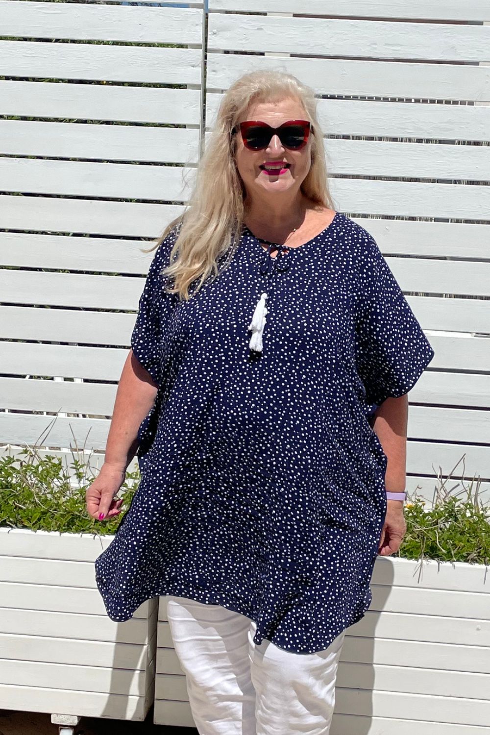 Mature age woman wearing the short kaftan dress in blue and white polka dot over white pants with planter boxes in background. 