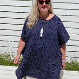 Mature age woman wearing the short kaftan dress in blue and white polka dot over white pants with planter boxes in background. 