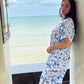 A woman wearing a blue and white floral print long kaftan dress stands in a doorway with a beach and ocean view in the background.