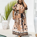 A curves models wearing size XL in long kaftan dress with short-sleeve in antique brass, black, and ghost white geometric graphic print design, styled with a black tie belt, set against a bright white wall with a potted palm poolside