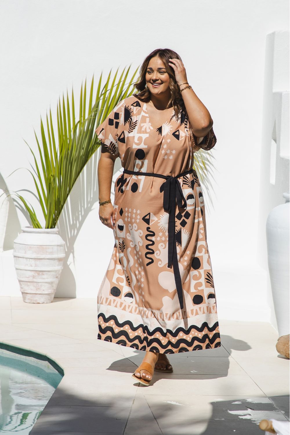 A curves models wearing size XL in long kaftan dress with short-sleeve in antique brass, black, and ghost white geometric graphic print design, styled with a black tie belt, set against a bright white wall with a potted palm poolside