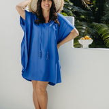 Alisha wearing the Lagoon Blue short kaftan dress in size SM with cream sandals and a woven hat beside tropical foliage.