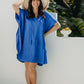 Alisha wearing the Lagoon Blue short kaftan dress in size SM with cream sandals and a woven hat beside tropical foliage.