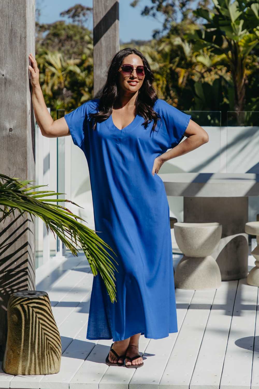 Front view of the Lagoon Blue long kaftan dress worn loose without a belt on a sunny deck with tropical foliage.