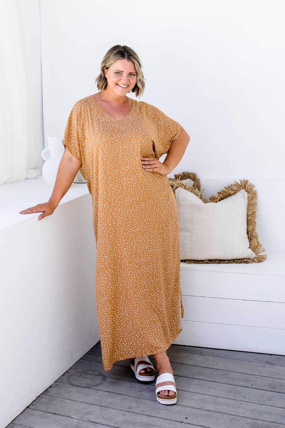 Amanda wearing ML Caramel Scatter dress, front-facing on a sunlit balcony beside a bench with cushions