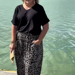 A woman stands outdoors by the water wearing a black short-sleeve top and loose black-and-white leopard-print pants, with sunglasses and a small tan clutch in hand.
