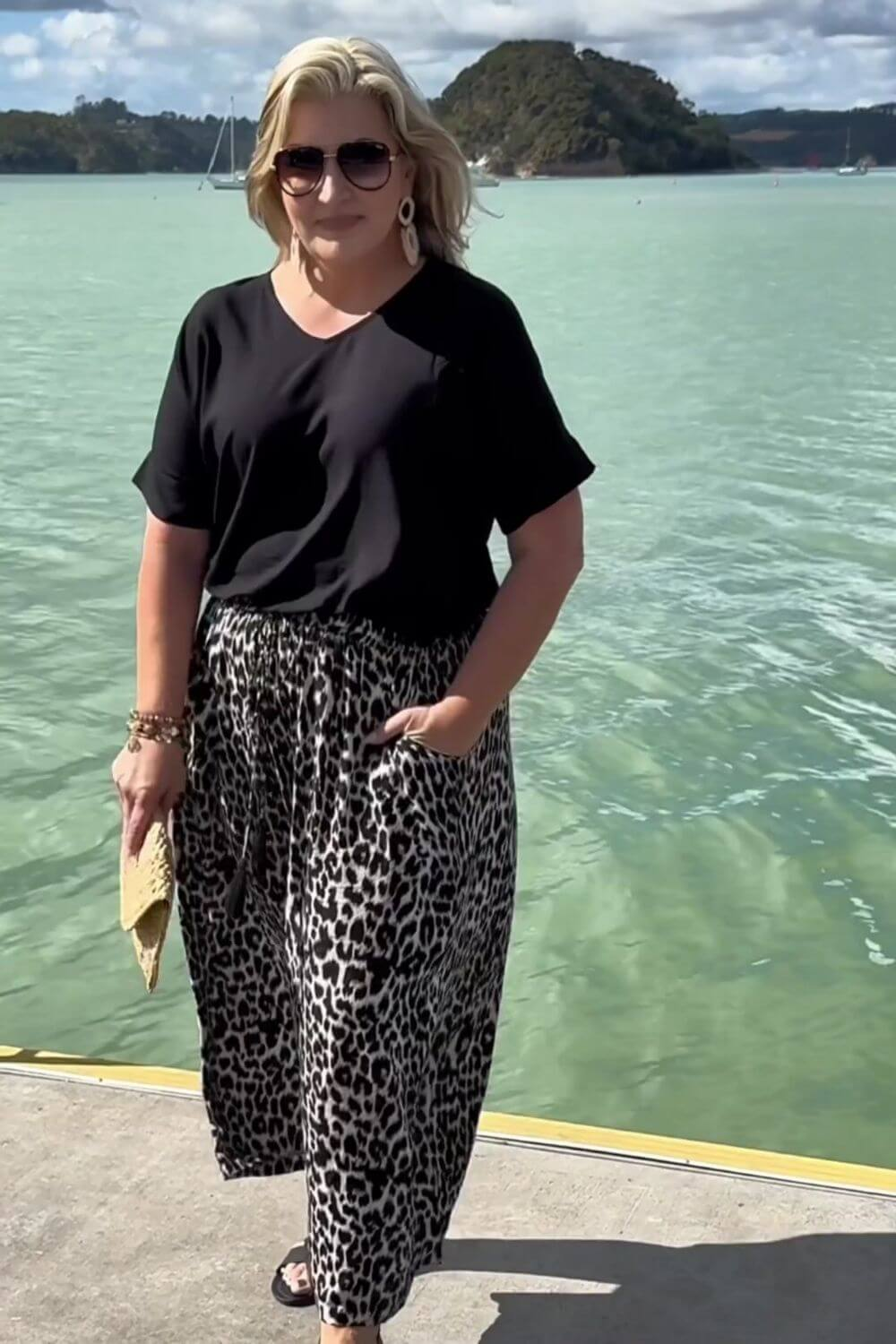 A woman stands outdoors by the water wearing a black short-sleeve top and loose black-and-white leopard-print pants, with sunglasses and a small tan clutch in hand.
