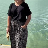 A woman stands outdoors by the water wearing a black short-sleeve top and loose black-and-white leopard-print pants, with sunglasses and a small tan clutch in hand.
