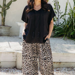 A woman wearing a black resort style kaftan top and wide-leg leopard-print pants, standing outdoor resort setting