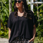 A woman wearing a black kimono sleeve top with tassels and leopard-print pants, standing outdoors in a tropical garden setting.