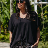 A woman wearing a black kimono sleeve top with tassels and leopard-print pants, standing outdoors in a tropical garden setting.