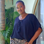 A woman wearing a relaxed fit black kaftan top with a tie neckline and wide leg leopard-print pants, standing in front of a tropical patio backdrop with palm leaves and a palm tree trunk.