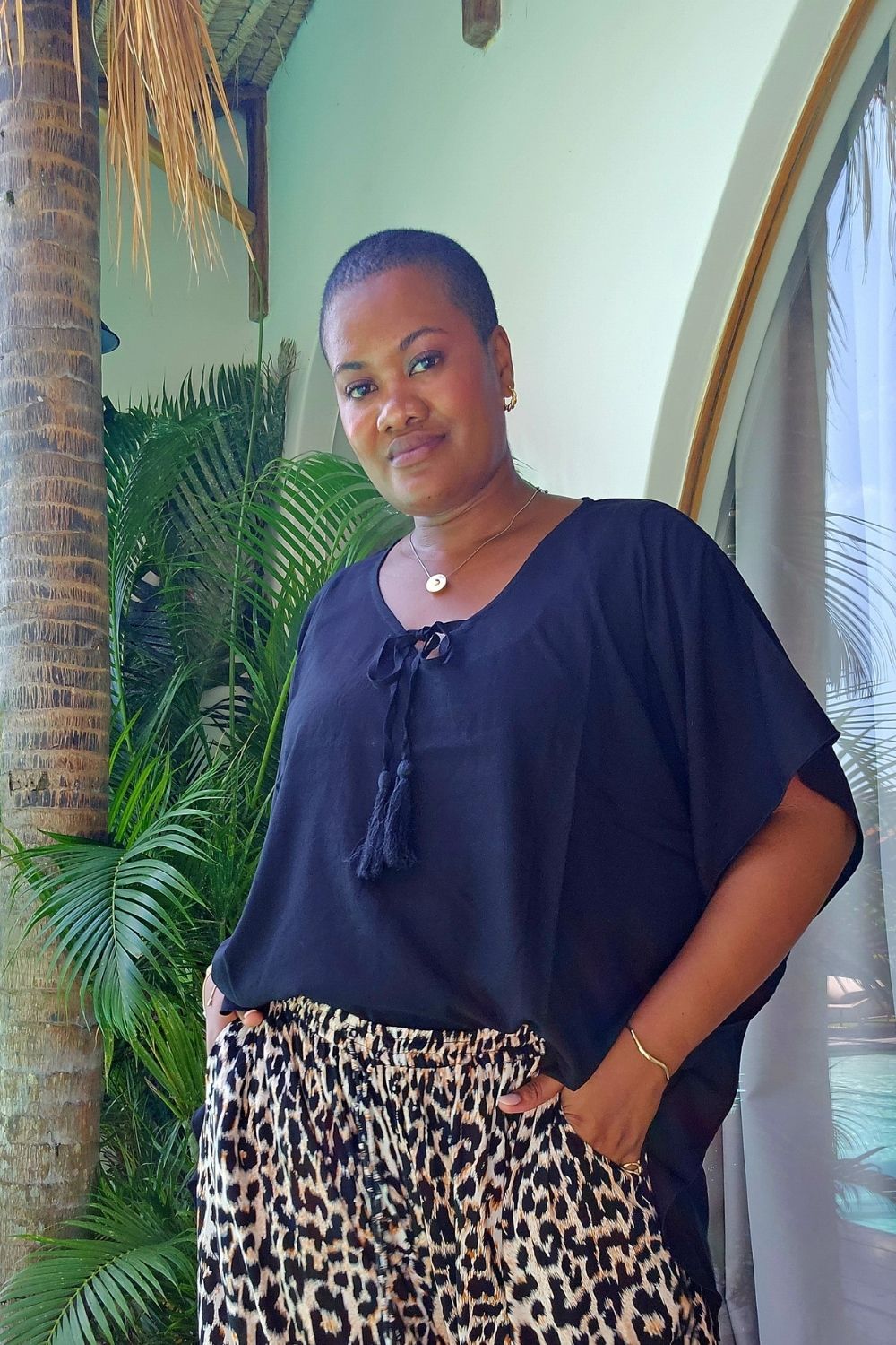 A woman wearing a relaxed fit black kaftan top with a tie neckline and wide leg leopard-print pants, standing in front of a tropical patio backdrop with palm leaves and a palm tree trunk.