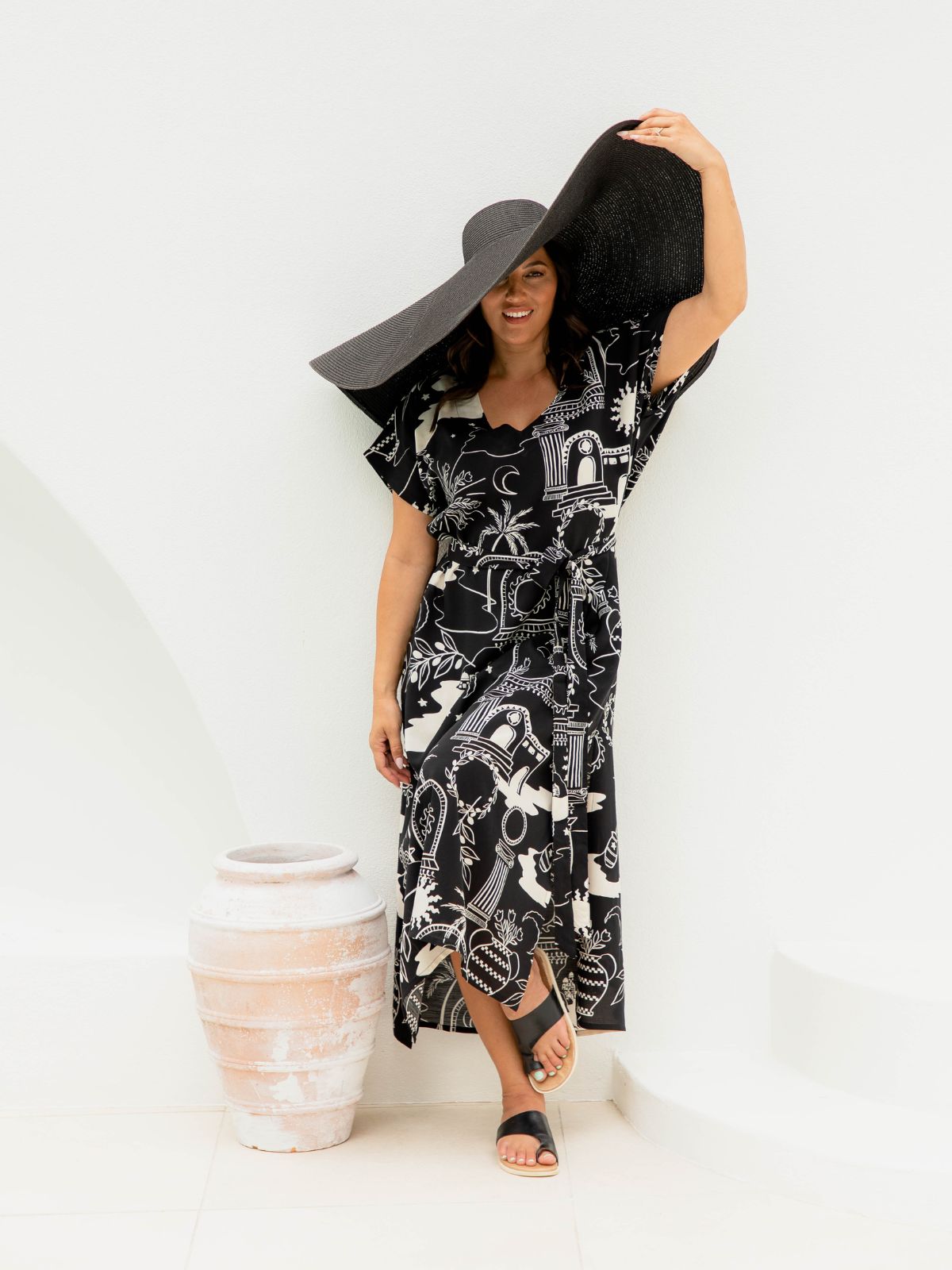 A woman modelling a black and cream graphic print long kaftan dress, wearing a large black sun hat and black sandals, standing in a bright white studio next to a rustic terracotta pot.