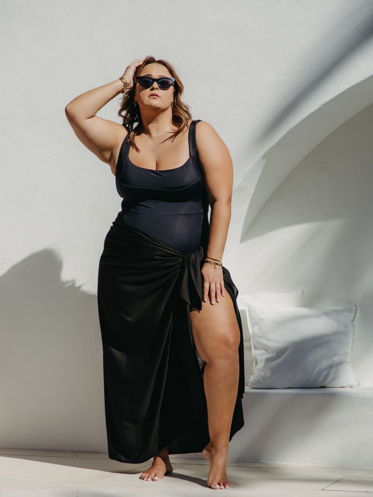 A woman modelling a black beach sarong skirt, standing in a sunlit white architectural setting poolside