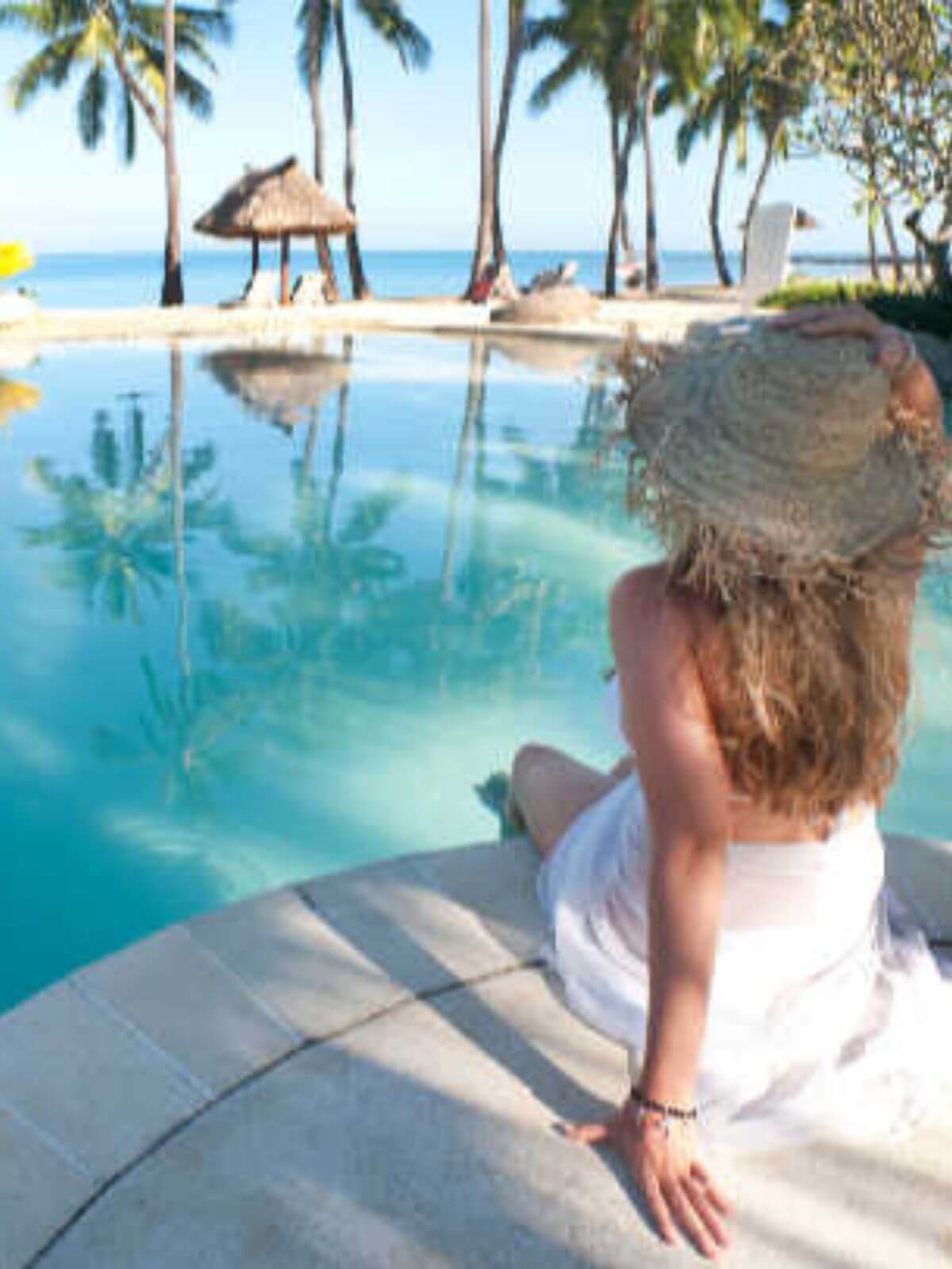 Woman sitting poolside wearing a white sarong overlooking a tropical beach with palm trees and ocean.