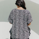 A woman viewed from behind wearing a black grey and white leopard print kimono sleeve kaftan tunic top and black pants, standing against a minimalist white wall outdoors