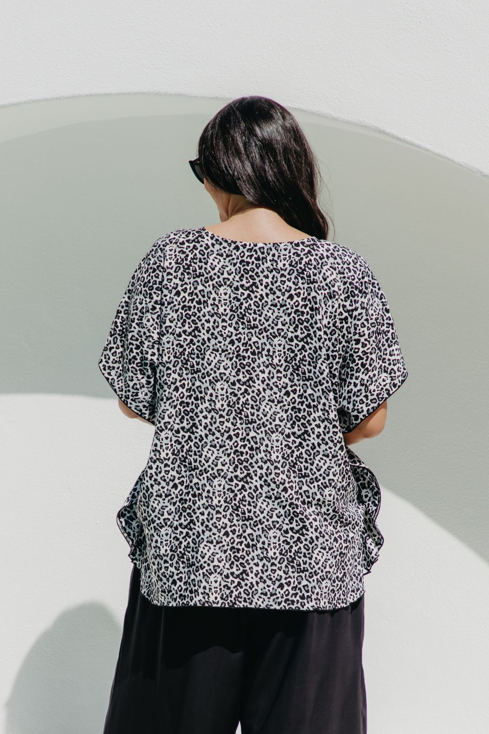 A woman viewed from behind wearing a black grey and white leopard print kimono sleeve kaftan tunic top and black pants, standing against a minimalist white wall outdoors