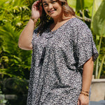 A woman wearing a black grey and white leopard print resort style kaftan top, standing outdoors in front of a lush green tropical background.