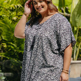 A woman wearing a black grey and white leopard print resort style kaftan top, standing outdoors in front of a lush green tropical background.