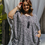 A woman wearing a black grey and white leopard print kaftan top and black cotton pants, posing in front of a white draped outdoor deck area.