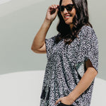 A woman wearing a black, white and grey leopard print summer kaftan top and black pants, posing outdoors against white arch 