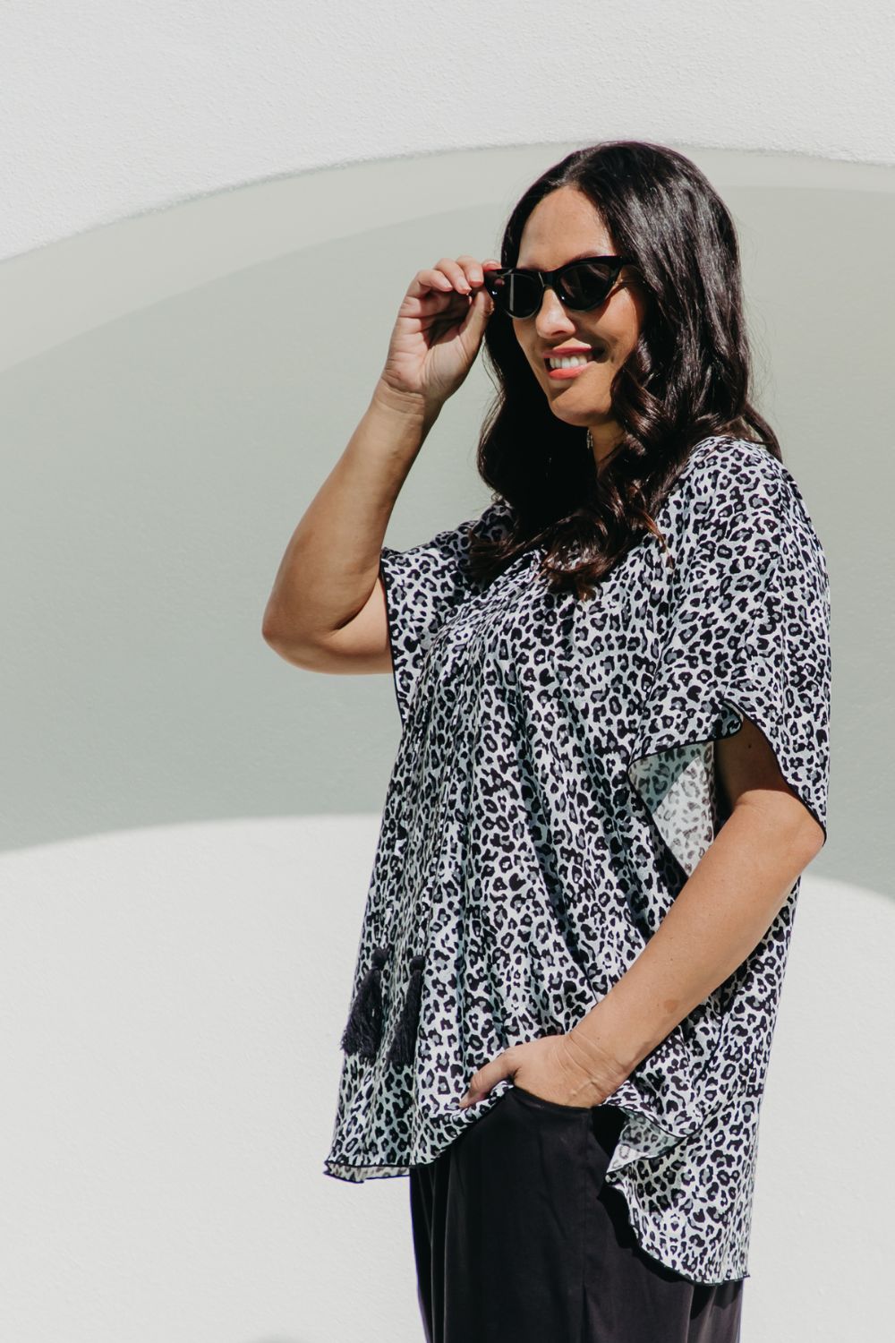 A woman wearing a black, white and grey leopard print summer kaftan top and black pants, posing outdoors against white arch 