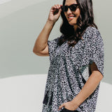 A woman wearing a black, white and grey leopard print summer kaftan top and black pants, posing outdoors against white arch 