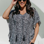 A woman wearing a black grey and white leopard print kimono sleeve kaftan top and black palazzo pants, posing outdoors against a white wall with a green arch.